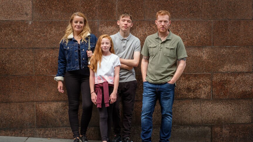 A woman, young girl, teenage boy, and man pose against an urban wall