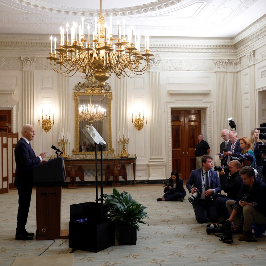 Joe Biden stands behind a lectern speaking with cameras and a group of journalists watching on