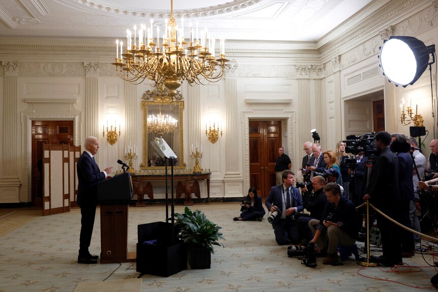 Joe Biden stands behind a lectern speaking with cameras and a group of journalists watching on