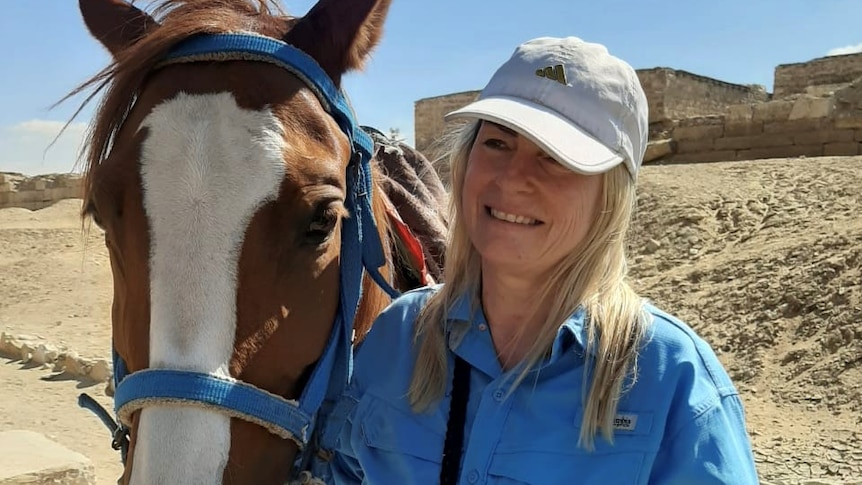 Jill Barton smiling next to a horse with the sandy Egyptian landscape behind her.