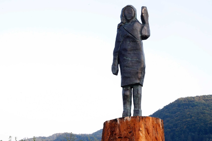 A bronze statue in the shape of a woman's figure stands atop a tree trunk. Her left hand is raised, mountains are the backdrop.