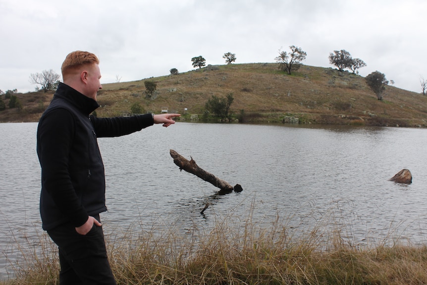 A man in a dark vest and dark trousers gesturing towards a dam.