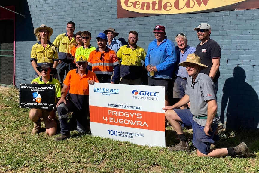 Large group of men standing next to a wall with a sign reading Fridgys for Eugowra.