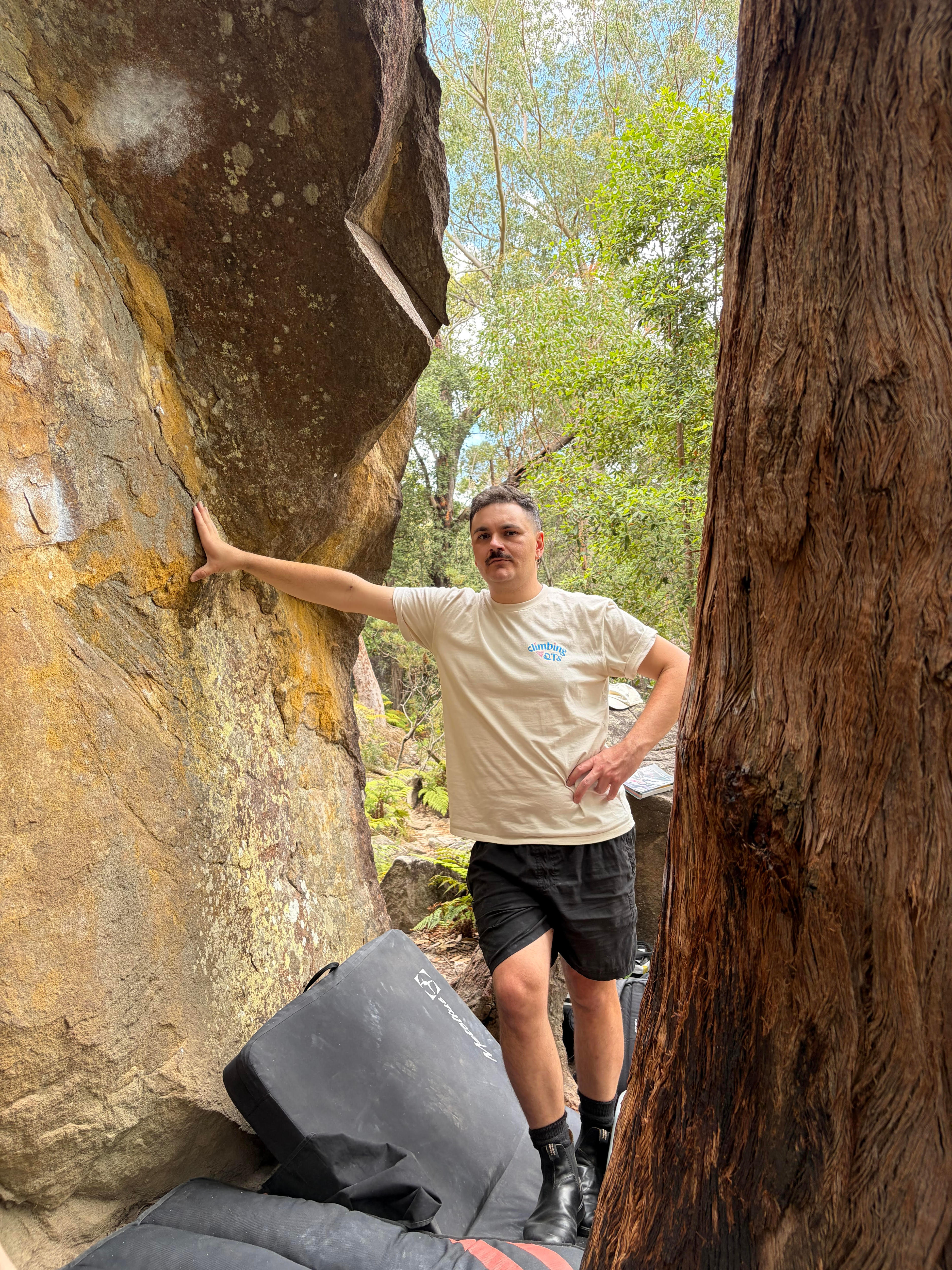A man with short, greying hair and a moustache leans against a rockface in a bushy area.