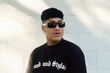 Jawsh 685 wears sunglasses and a black t-shirt, he is standing against a white brick wall.