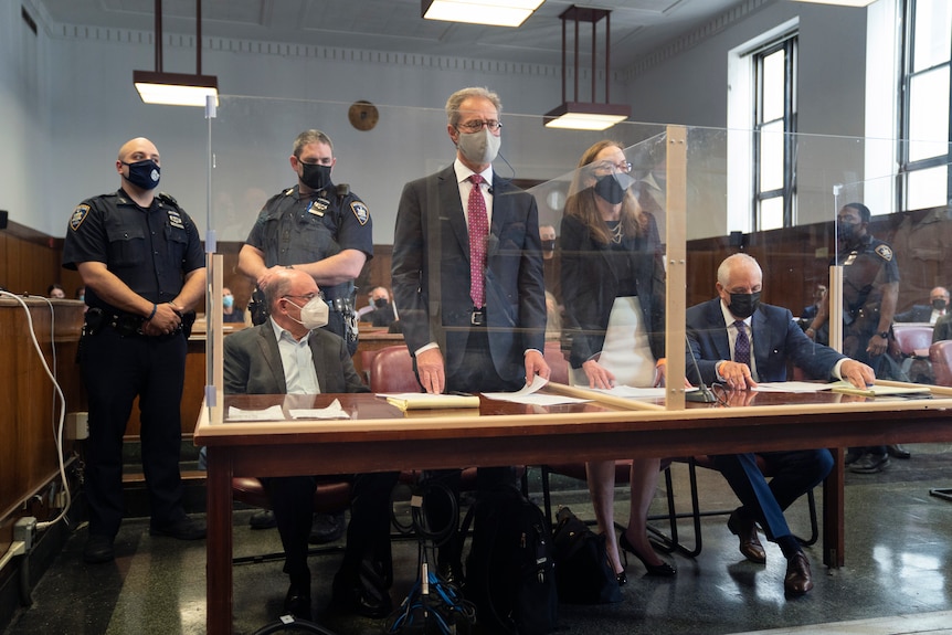 A man in a mask sits next to several lawyers behind a clear screen in a court room.