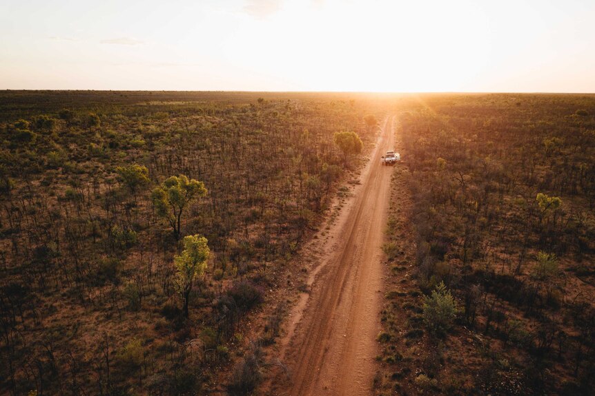 Aerial shot of a car stopped by the side of a dirt road in the desert at sunset.