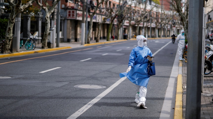 A worker in full PPE walks down an empty street lined with trees and buildings.