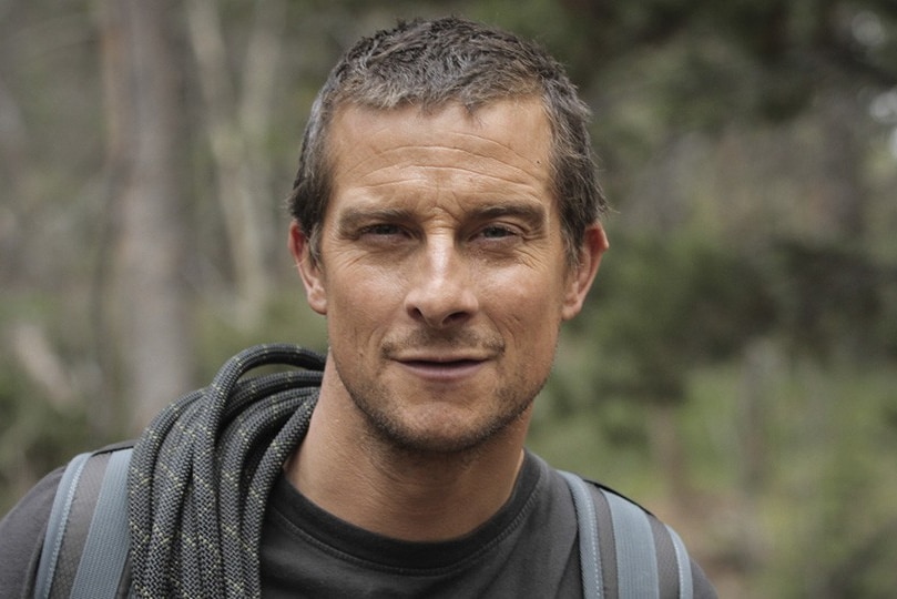Survivalist Bear Grylls in the wilderness with a coil of rope over his shoulder and a hawk-like expression on his face.