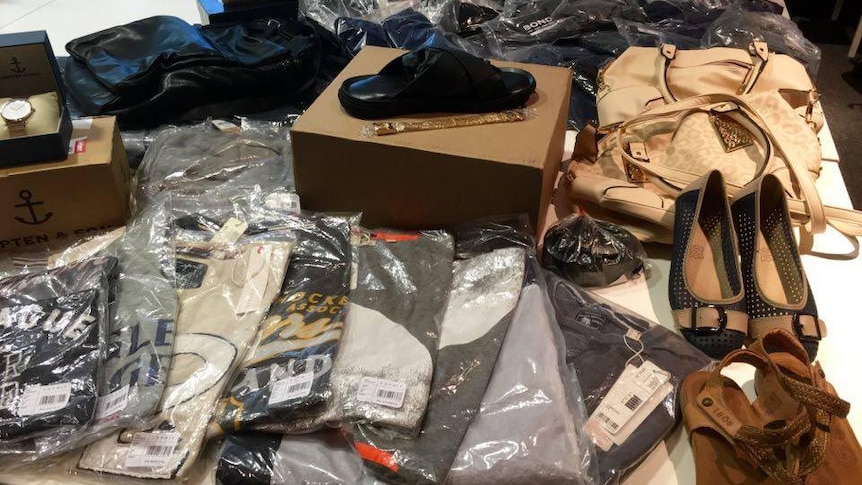 A group of items on a table including t-shirts in plastic bags, shoes, bags and a watch.