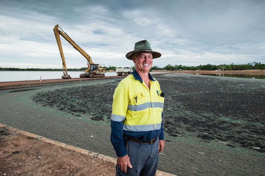 man hi-viz standing before sewage pond with excavator in background moody cloudy sky