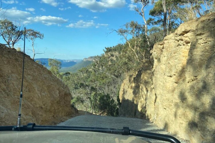 Steep unsealed road falls away between sandstone walls, photo through front window of a 4WD