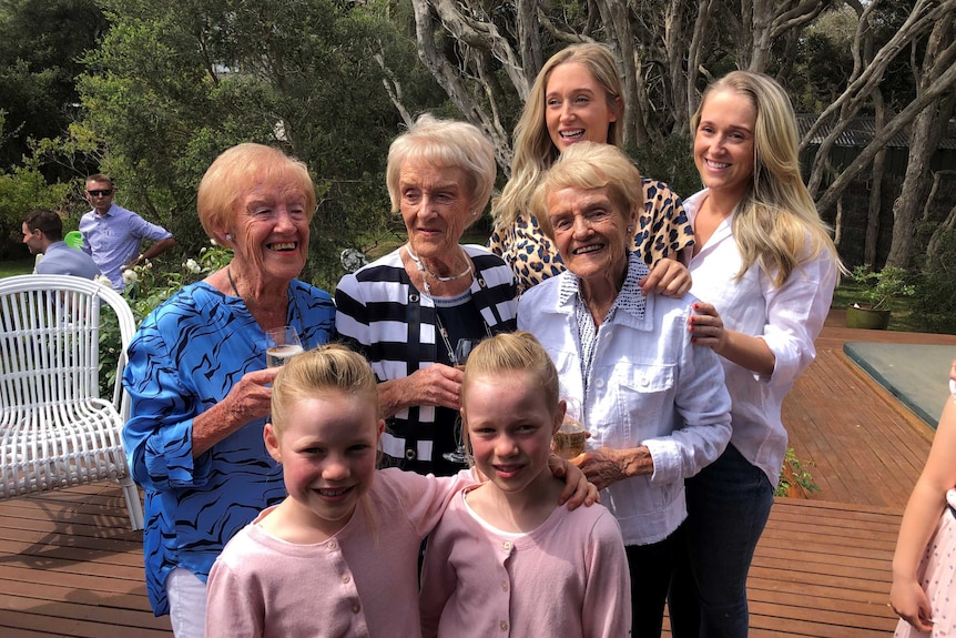 Three smiling 90-year-old women stand with two women in their 20s and two girls