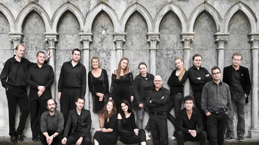 An ensemble of singers, dressed in grey and black, standing in front of grey cement gothic arches.