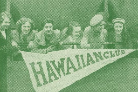 Black and white photo with green tinge, of women leaning out over a ledge smiling widely, with 'Hawaiian Club' sign below them.