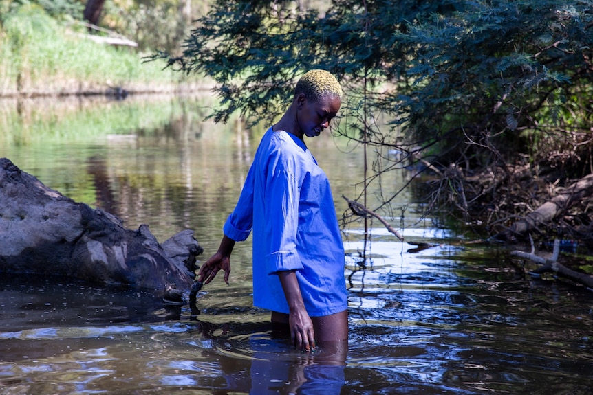 A still of an African woman in a button-down shirt looking down as she wades into a creek, the water at her thighs