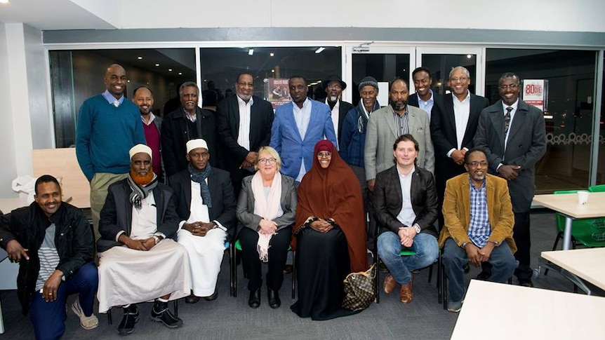 A group of 17 Somali community leaders, some sitting some standing, pose with Jenny Macklin, in a community centre hall.