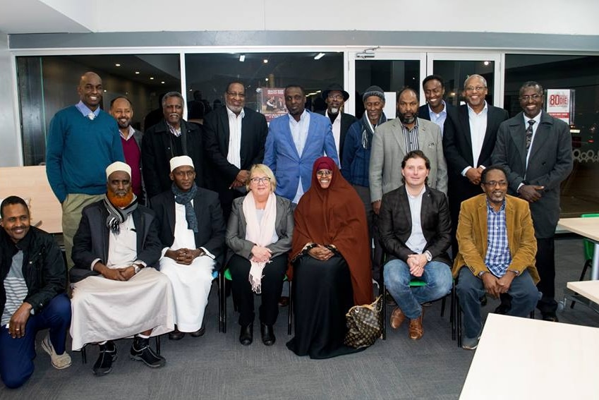 A group of 17 Somali community leaders, some sitting some standing, pose with Jenny Macklin, in a community centre hall.