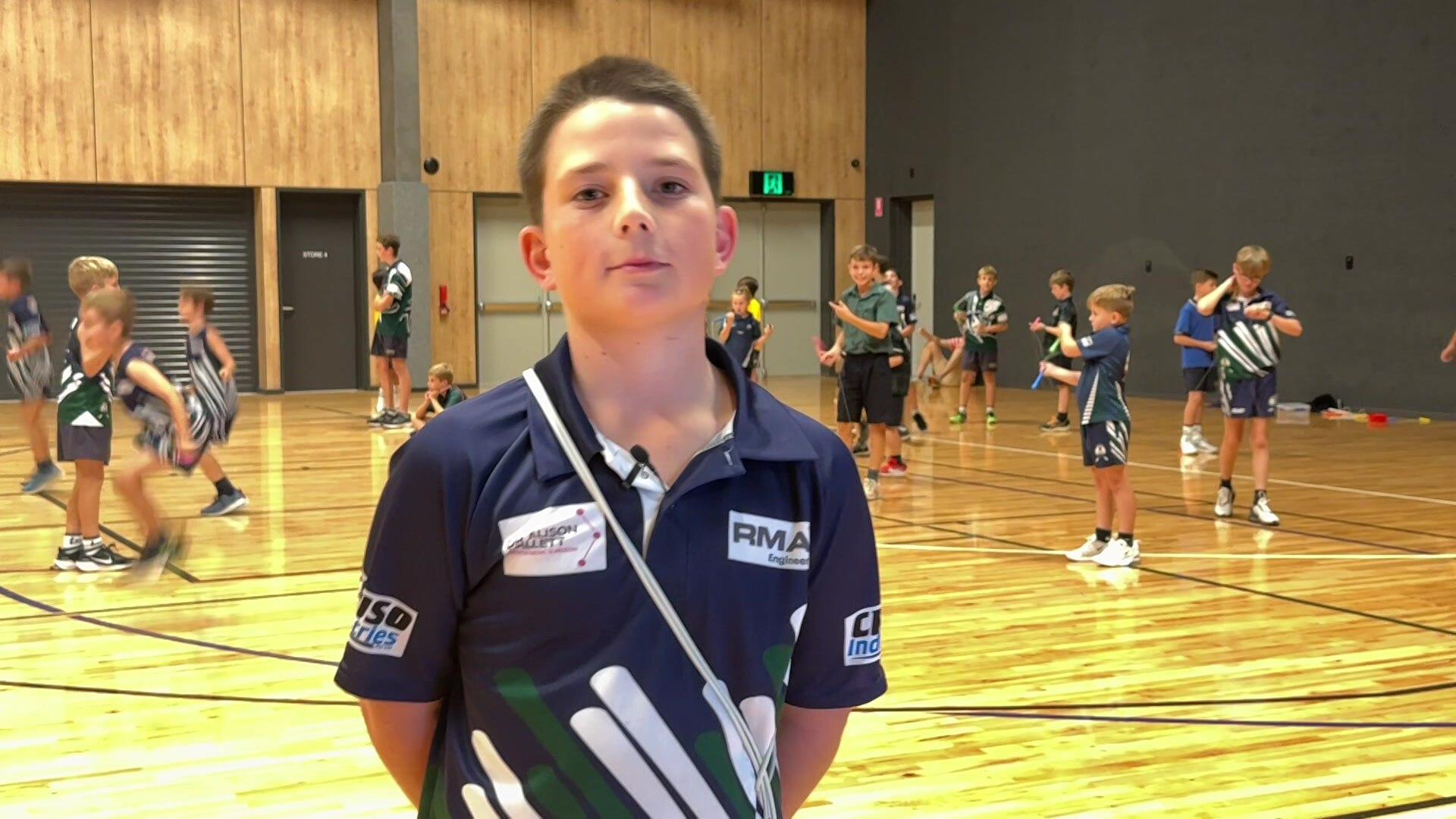 A young boy standing in a school gym with students skipping behind him.
