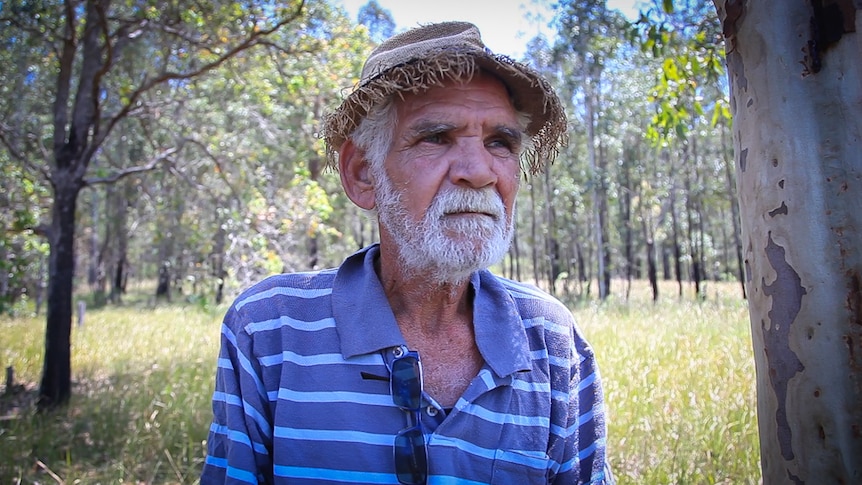 Older Indigenous man wearing a straw hat is standing in bushland on a farm.