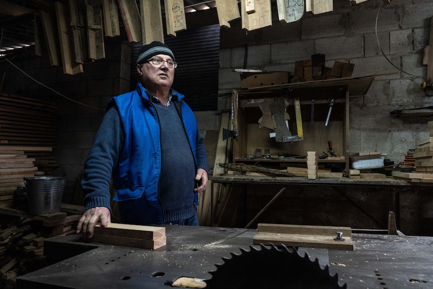 An older man wearing a blue puffer vest stands at a workshop bench, with timber scraps and saws
