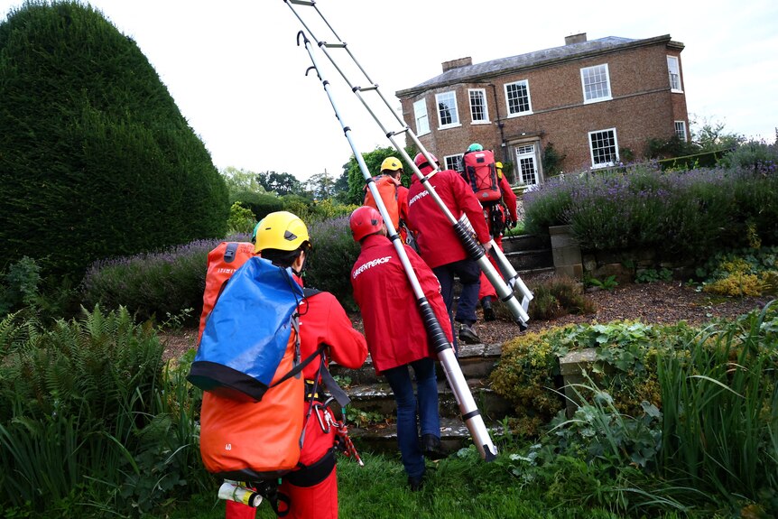 Five people wearing orange suits and carrying ladders and climbing gear walk through garden towards big house