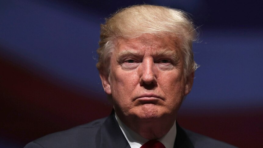 Donald Trump stares at the camera with a neutral expression in a relatively close photo. Behind him the background is darkened.