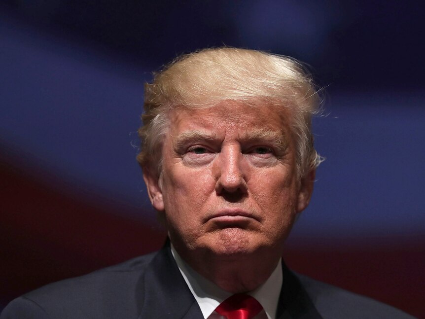 Donald Trump stares at the camera with a neutral expression in a relatively close photo. Behind him the background is darkened.