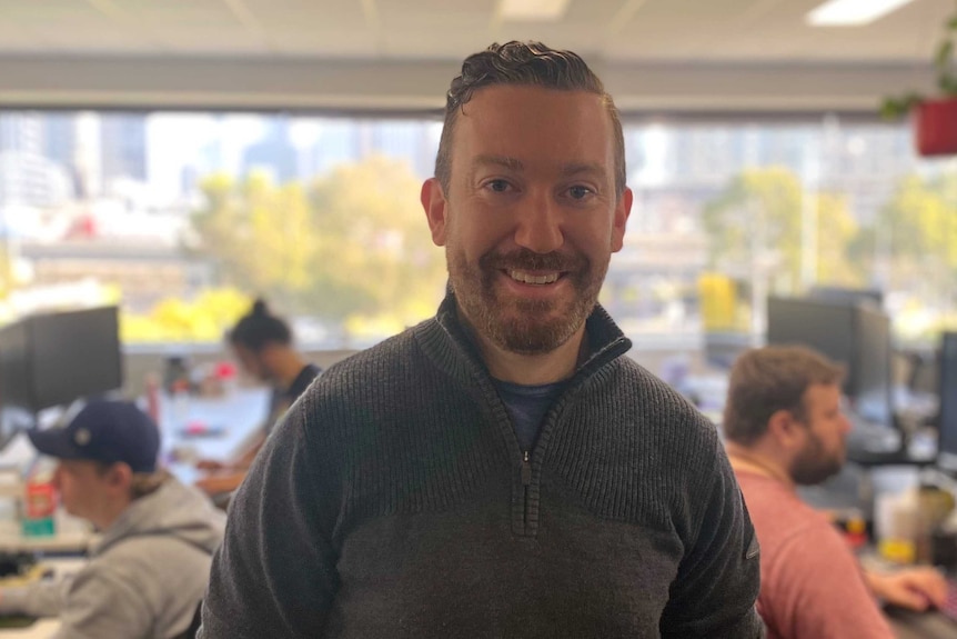 A man with a beard and mustache smiling in a room full of people on computers.