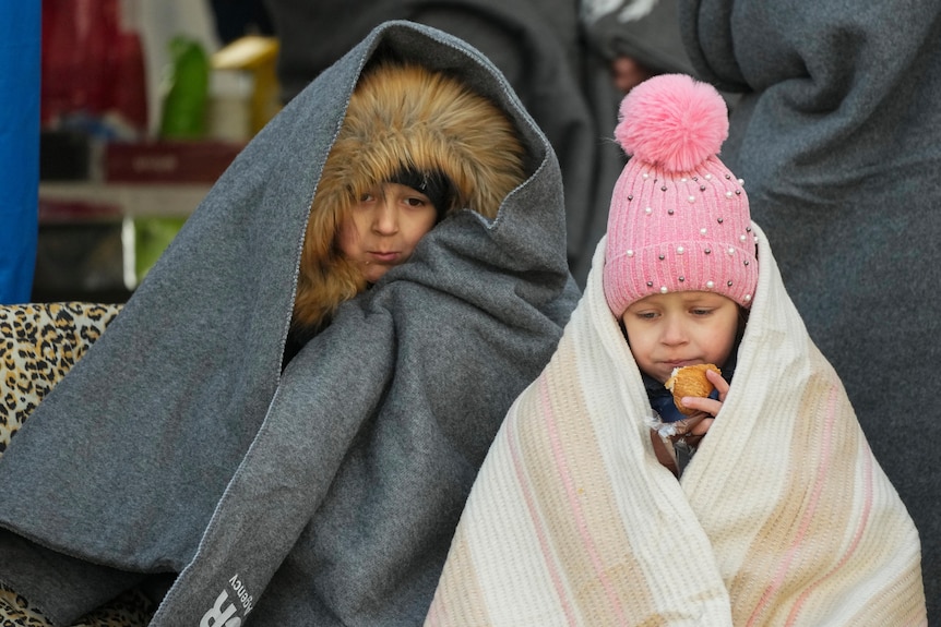 Two children are wrapped in blankets wearing beanies while eating.