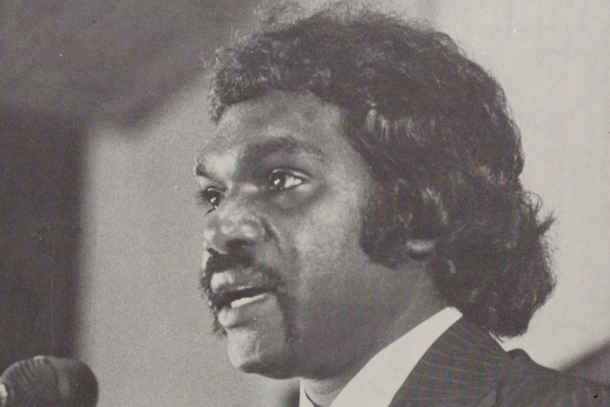 A black and white, historical of Gumatj leader Galarrwuy Yunupingu as a young man, dressed in a suit and speaking at an event.