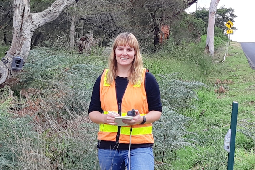 Woman stands in bush beside the side of the road wearing a hi-vis vest. She is smiling.