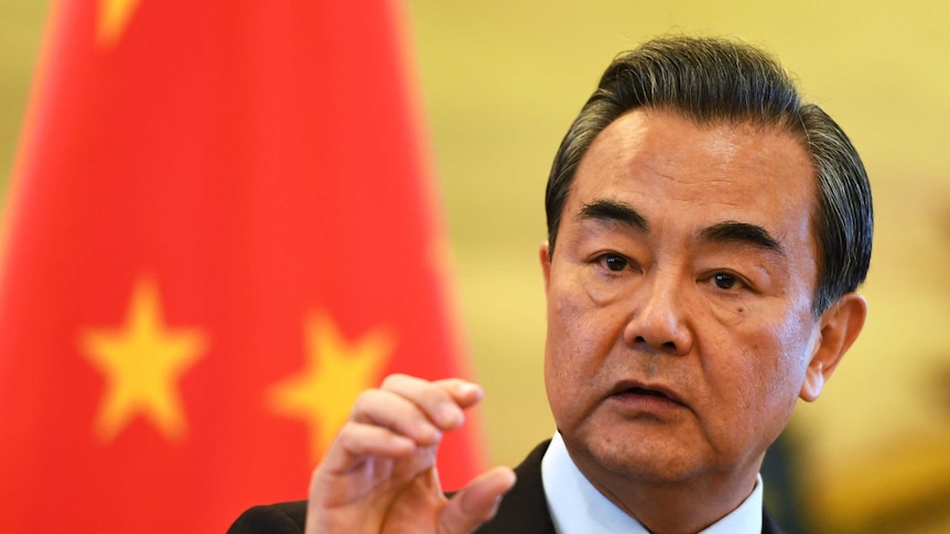 An older Asian man looking to his right with his hand raised while he stands in front of the Chinese flag