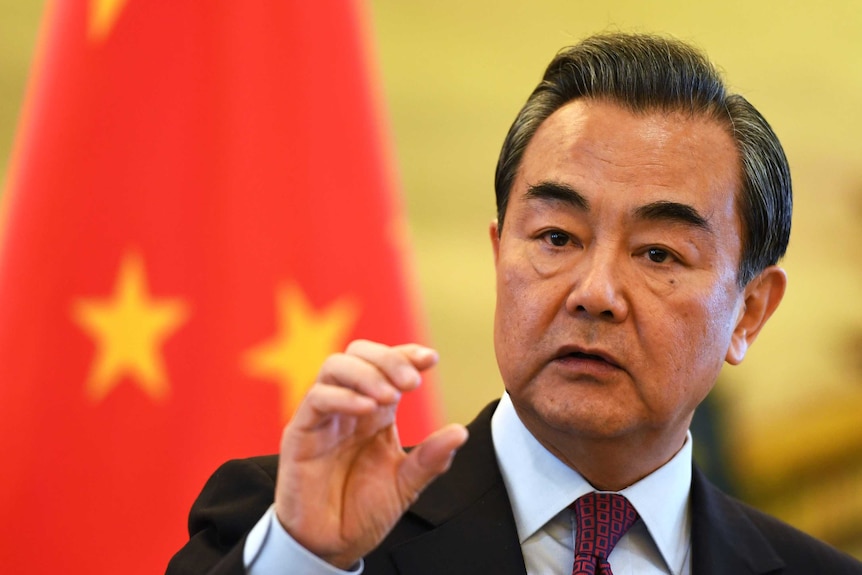 An older Asian man looking to his right with his hand raised while he stands in front of the Chinese flag