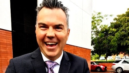 A middle-aged man in a suit and tie laughs while standing on the pavement near a building.