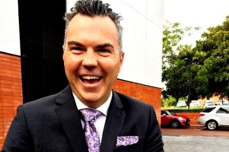 A middle-aged man in a suit and tie laughs while standing on the pavement near a building.