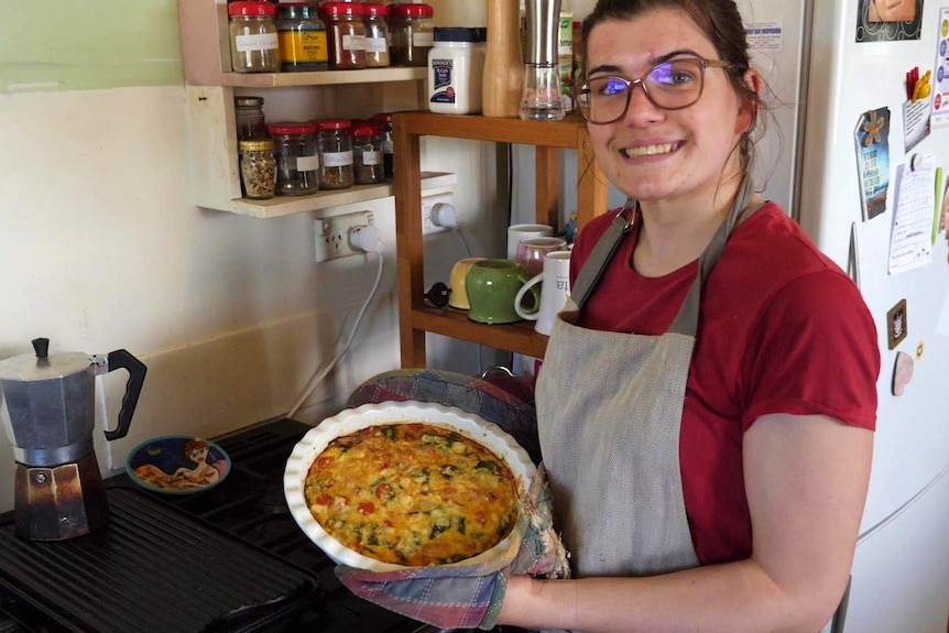 Teenage girl with glasses on right holding a plate of quiche up in kitchen