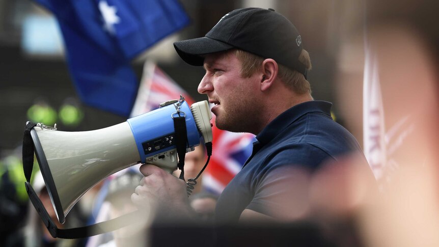 Blair Cottrell holds a megaphone to his mouth. Many Australian flags are visible in the background.