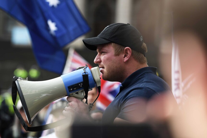 Blair Cottrell holds a megaphone to his mouth. Many Australian flags are visible in the background.