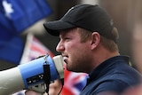 Blair Cottrell holds a megaphone to his mouth. Many Australian flags are visible in the background.