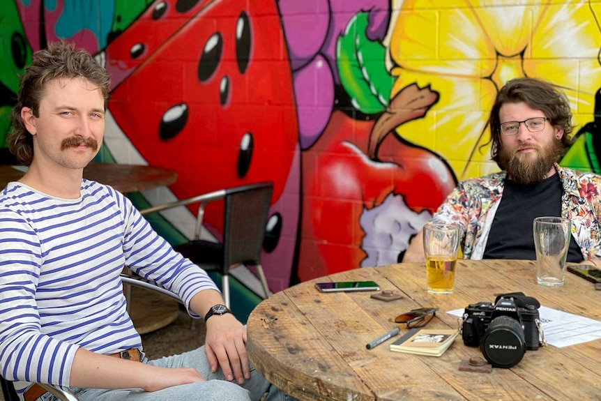 Two men sit at a table drinking beer with fruit street art behind them.