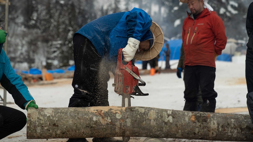 A Japanese man uses a chainsaw to cut up wood for the fire festival while his friend looks on.