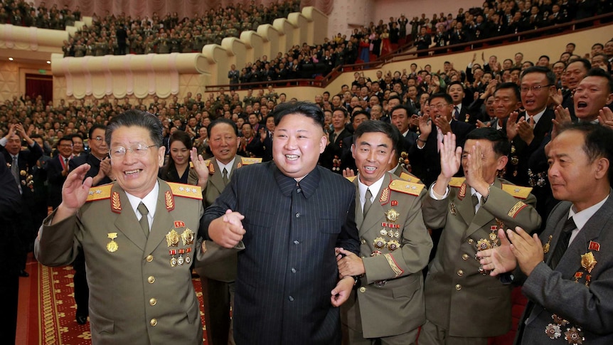 Kim Jong-un smiles in front of a huge crowd in a concert hall. They are celebrating the country's nuclear scientists.