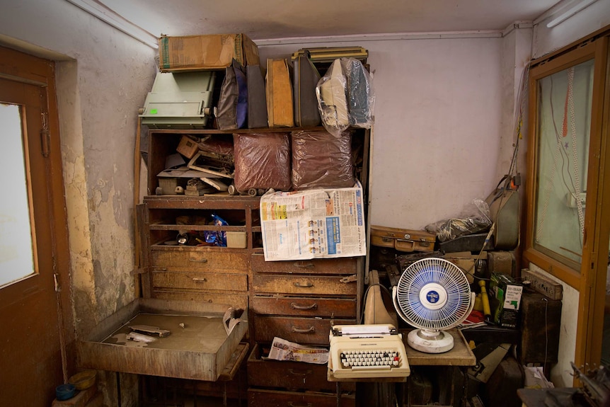 An old worn set of wooden drawers with shelved packed with bags and objects, in a small room
