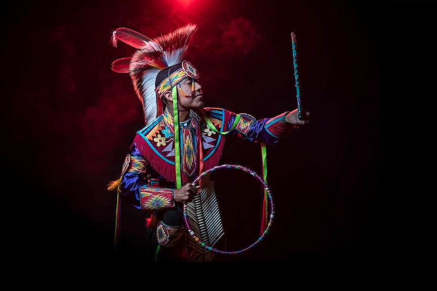 In front of red smoke a man in bright coloured Native American regalia holds hoop and looks at self in handheld mirror.