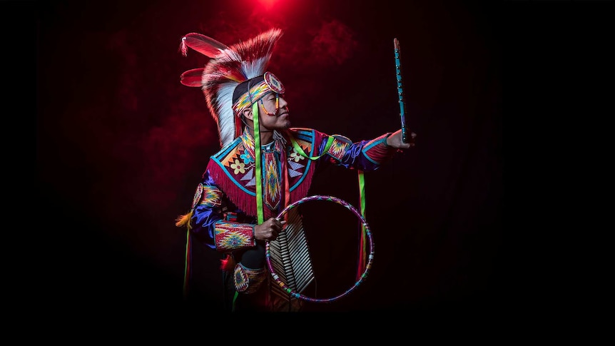 In front of red smoke a man in bright coloured Native American regalia holds hoop and looks at self in handheld mirror.