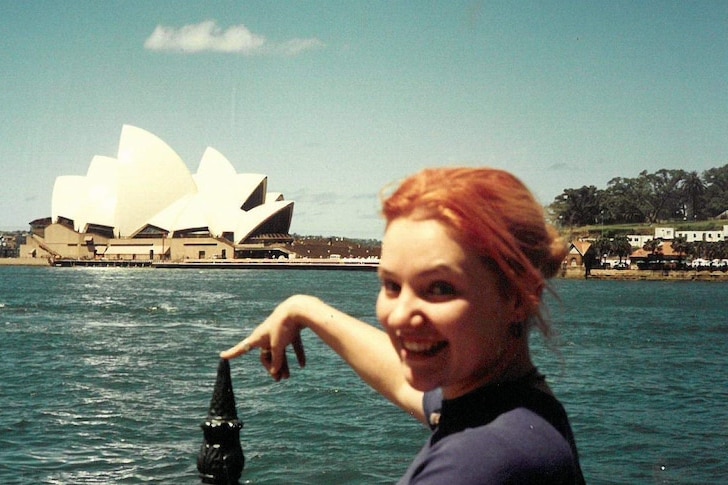 Photo of Jacinta Parsons in her younger years, with orange hair, smiling widely with Sydney Opera House in the background.