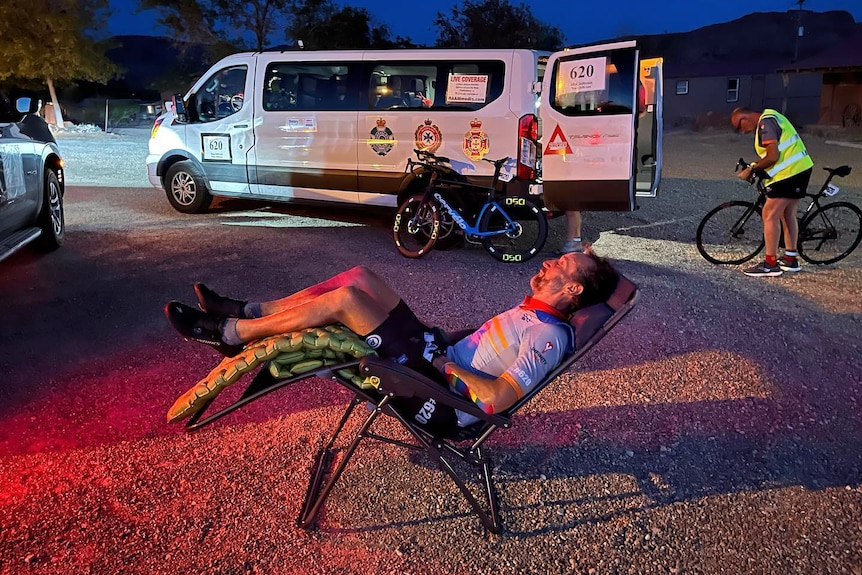 A cyclist sleeps in a reclining chair in front of a support van