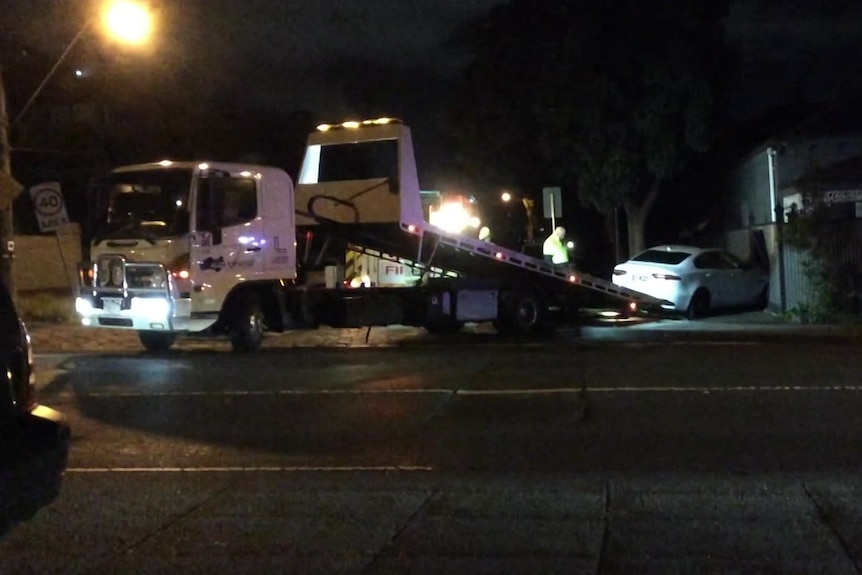 A night scene of a tow truck next to a white car crashed into a fence.