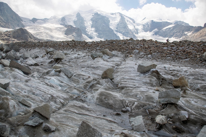 A hiker in the background walks over a glacier. Rocks and ice can be seen in the foreground. 